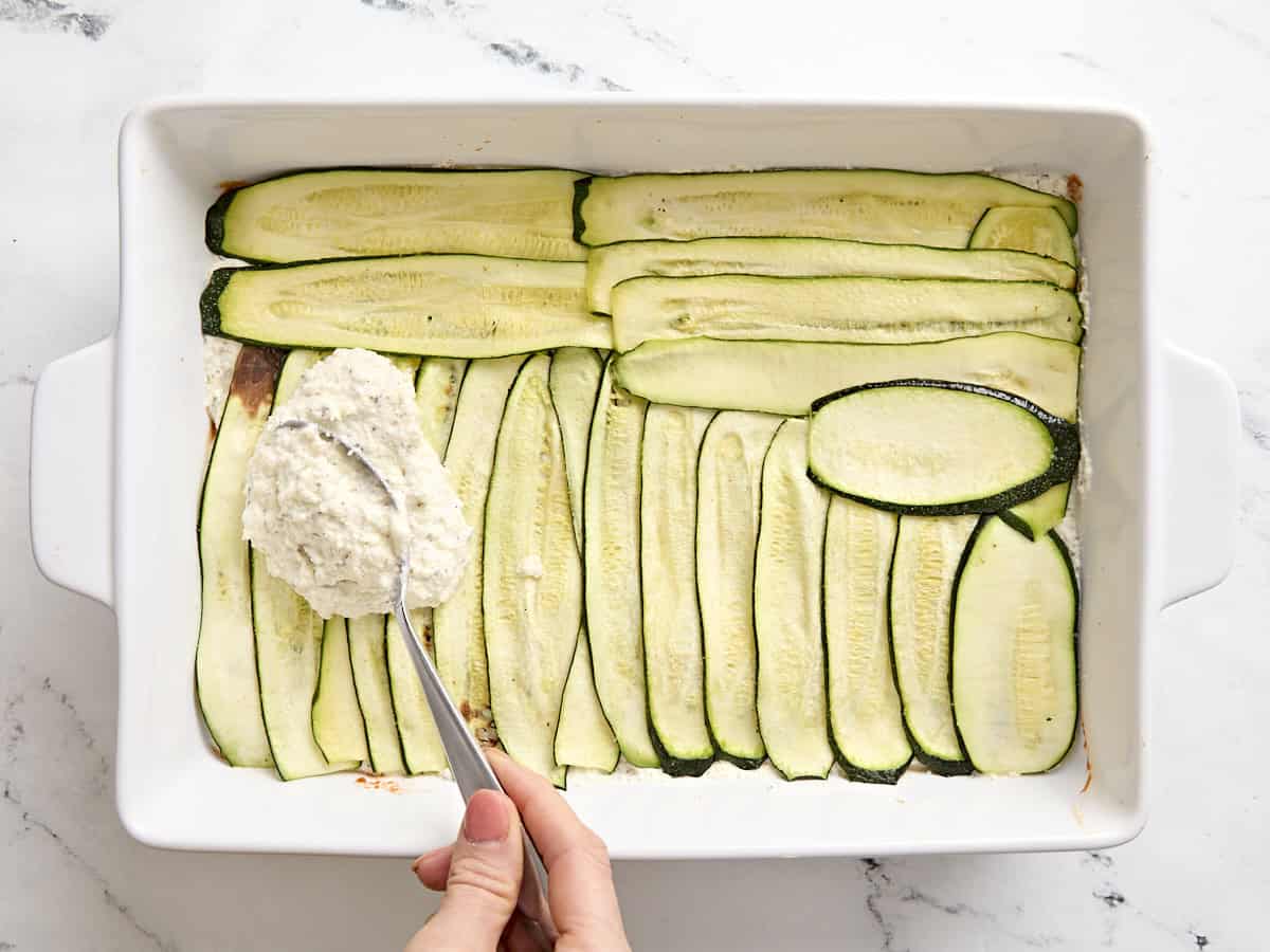 A spoon adding cheese sauce to zucchini slices in a baking dish for zucchini lasagna