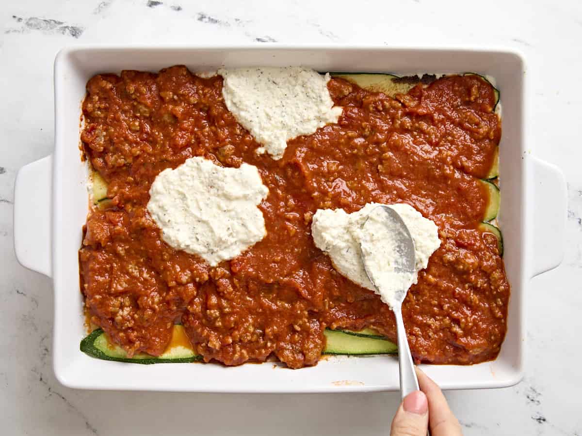 A spoon dolloping cheese sauce onto zucchini lasagna