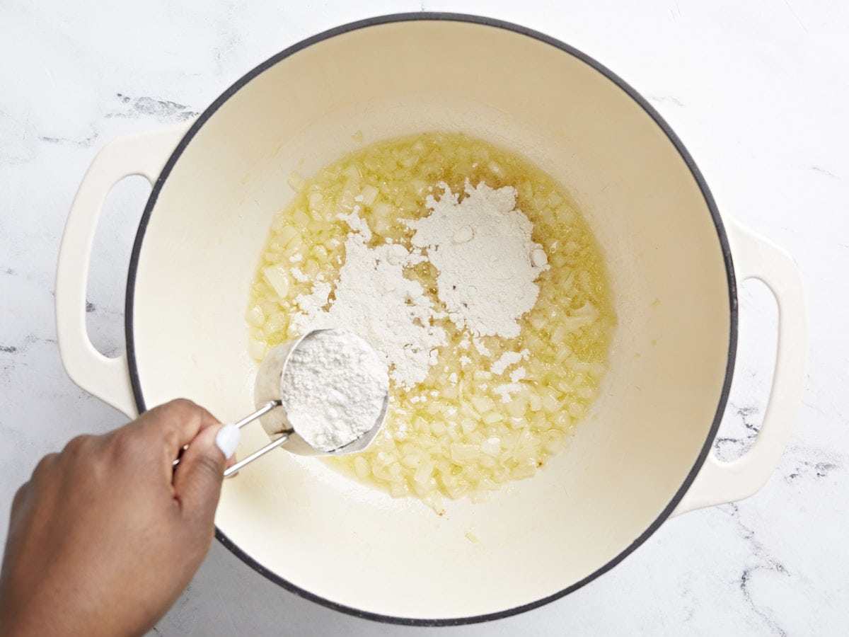 Overhead view of sauteed onions and garlic in a large dutch oven pot with flour being sprinkled over the top.