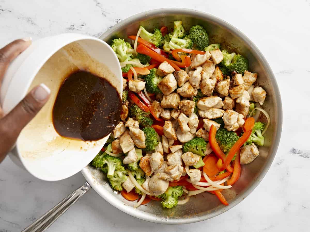 Cooked chicken and stir fry sauce being added to vegetables in a skillet.