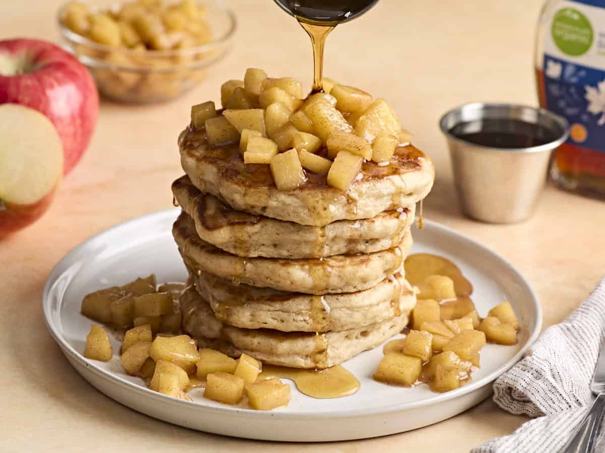 A stack of apple cinnamon pancakes with maple syrup being drizzled over top.