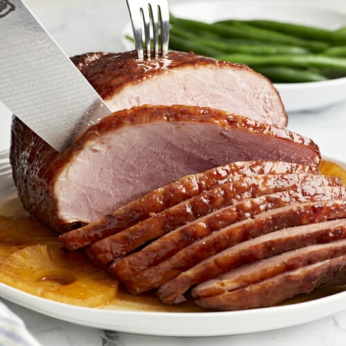 slicing a baked ham on a white plate with pineapple rings.