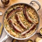 Bratwurst and sauerkraut in a skillet with beer, potatoes, and mustard on the sides