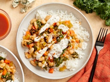 overhead view of buffalo beans and greens over white rice on a white plate with white sauce drizzled over top.