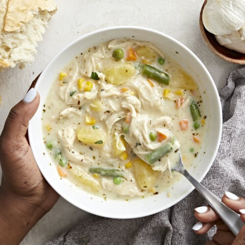 Overhead view of a serving bowl of chicken pot pie soup with one hand holding the bowl and the other hand holding a spoon .