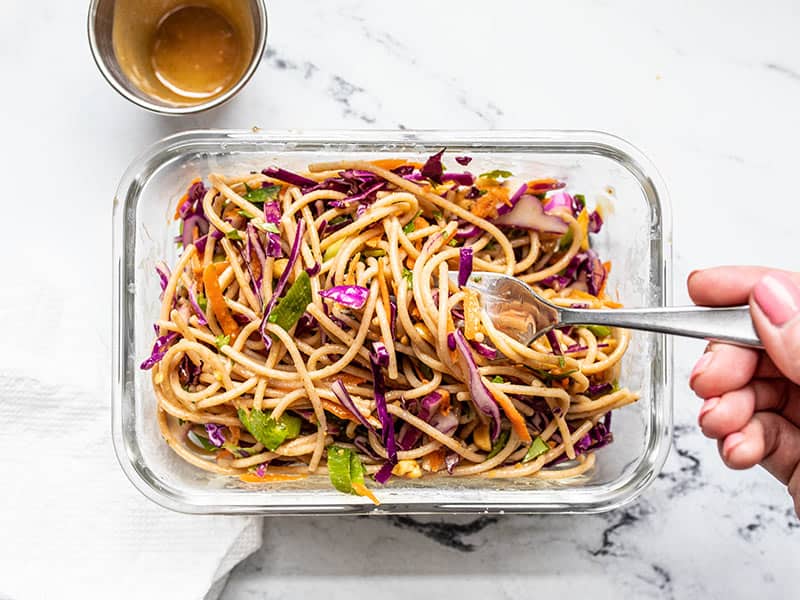 A meal prep container of Cold Peanut Noodle Salad being eaten with a fork, the empty dressing container on the side.