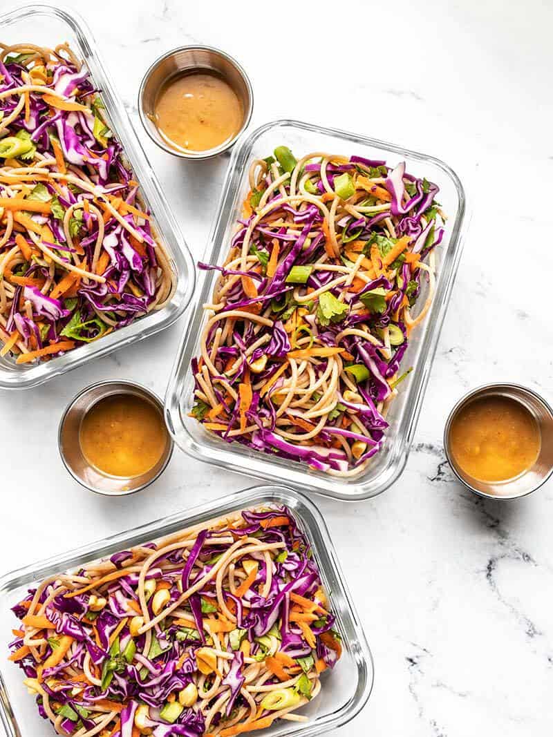 Three glass meal prep containers full of cold peanut noodle salad, dressing containers on the side.