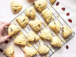 A hand taking one Cranberry Orange Cream Scone from the batch on a cooling rack.