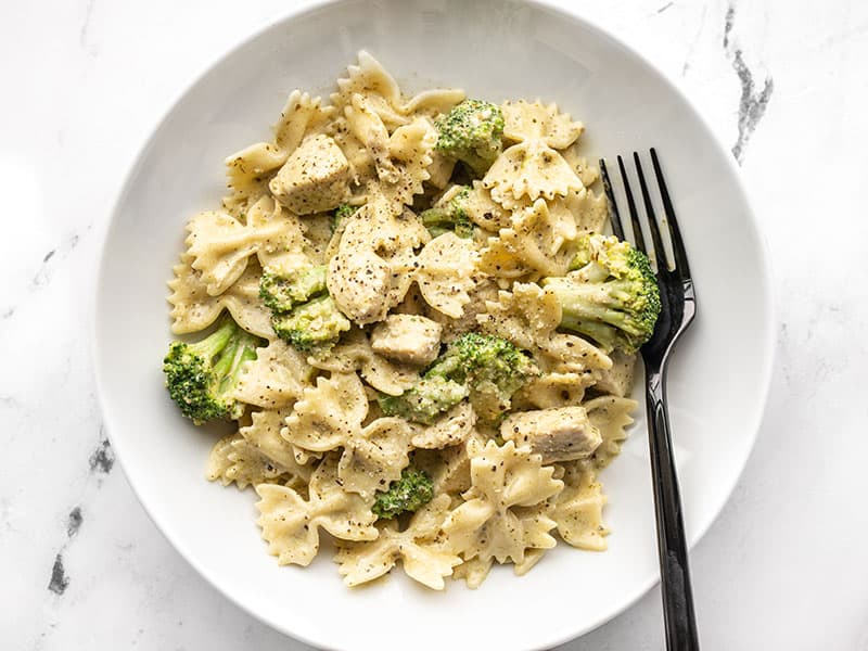 A bowl full of creamy pesto pasta with chicken and broccoli, a black fork on the side