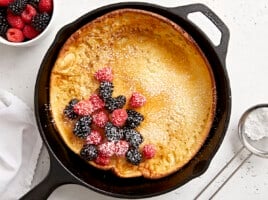 Overhead view of a dutch baby in a cast iron skillet with fresh berries in the center and maple syrup on the side.