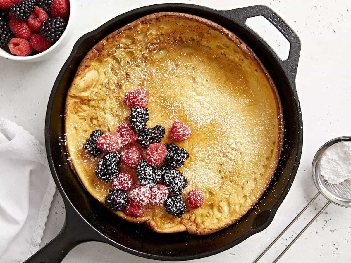 Overhead view of a dutch baby in a cast iron skillet with fresh berries in the center and maple syrup on the side.