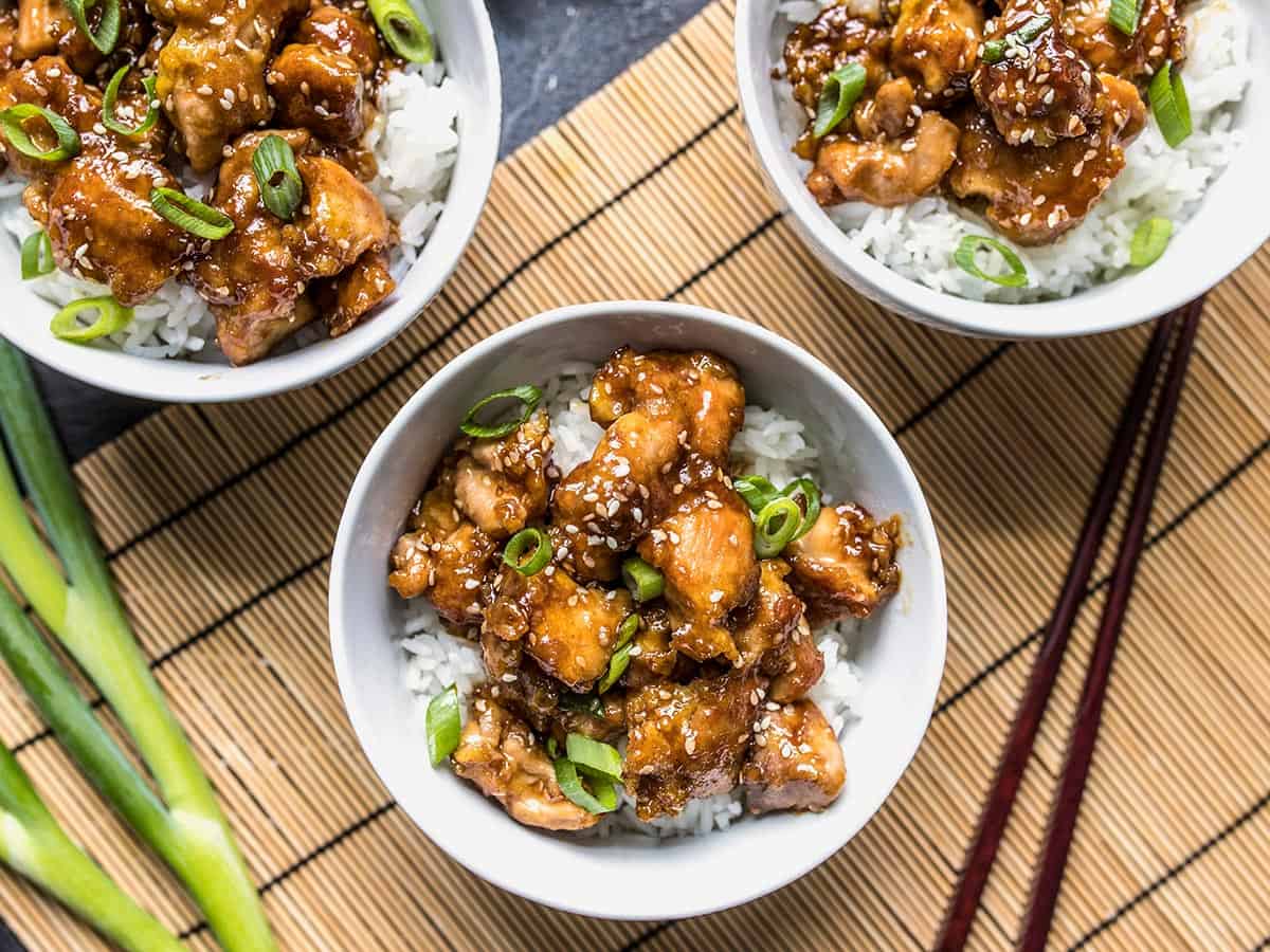 Overhead view of three bowls of sesame chicken with white rice and green onion. 