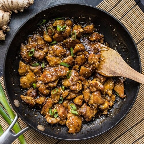 Overhead view of a frying pan full of sesame chicken with a wooden spatula.