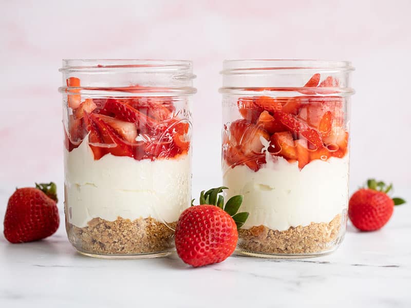 Two jars of no bake strawberry cheesecake side by side with whole strawberries on the side