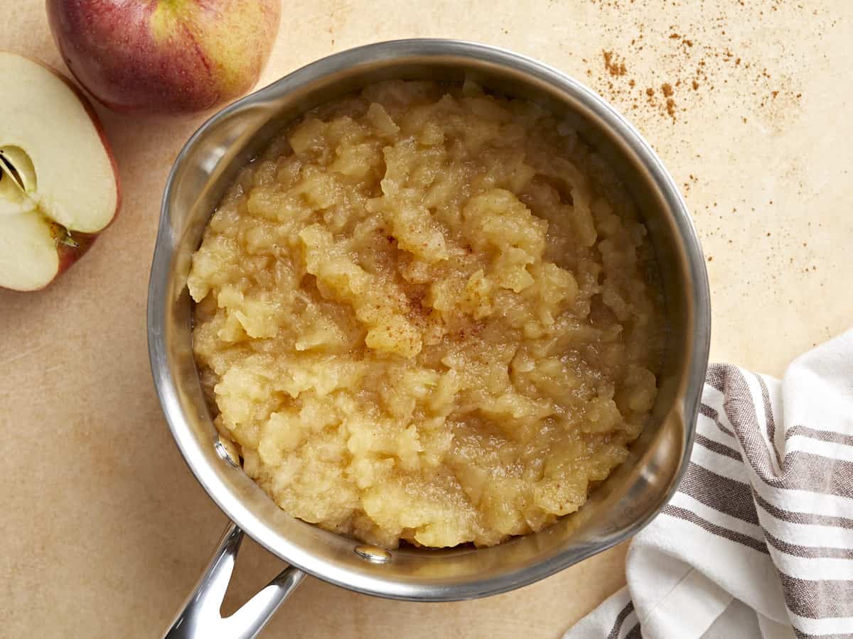 Homemade applesauce in a small saucepan with apples and a napkin on the side.