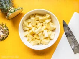 A bowl of fresh pineapple chunks next to a cutting board, chef's knife, and a pineapple top.