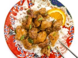 CLose up overhead view of orange chicken on a plate with rice, green onion, and orange slices.