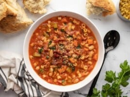 A white serving bowl of pasta e fagioli soup topped with bacon, parsley and parmesan cheese, and surrounded by torn bread, uncooked pasta, a black serving spoon, italian parsley and a decorative blue and white dish cloth.