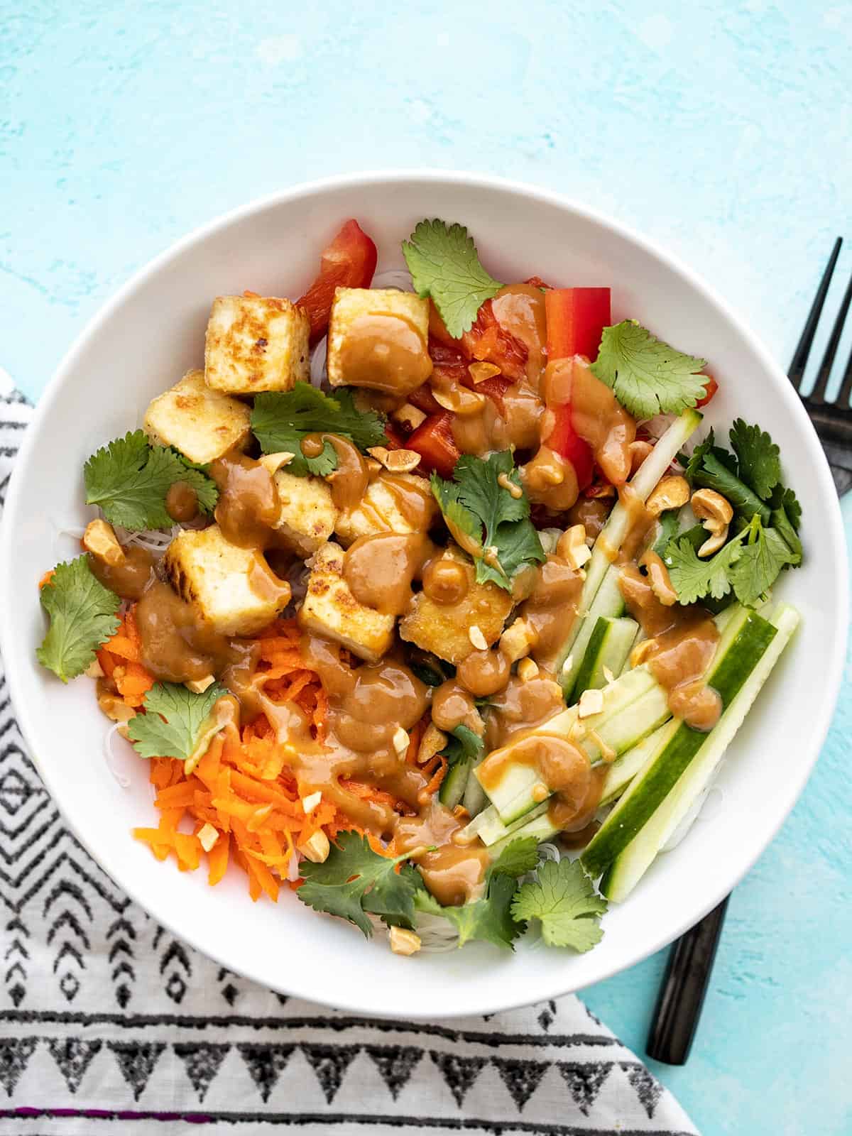 Overhead view of a peanut tofu noodle bowl on a blue background and a fork on the side