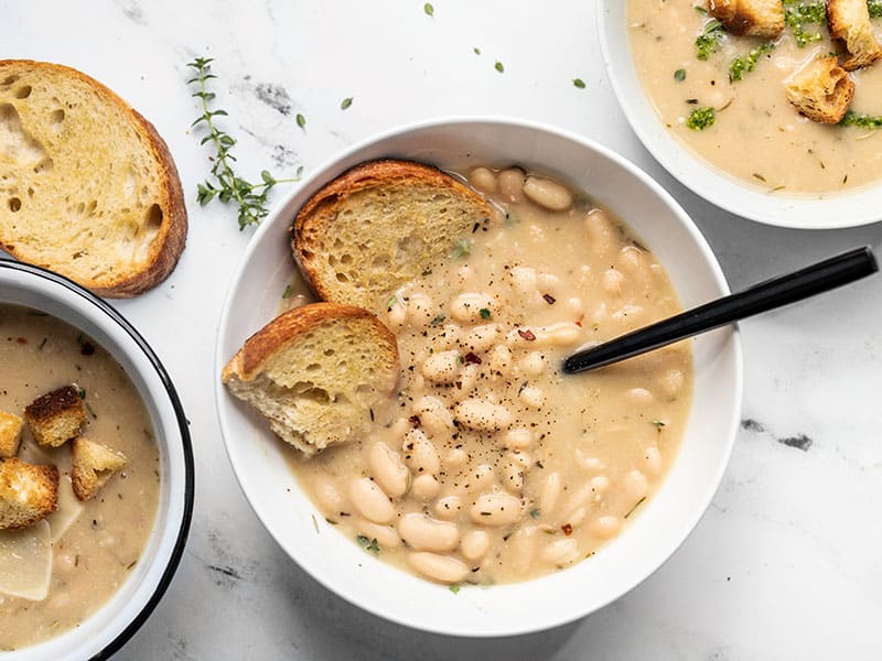 Three bowls of rosemary garlic white bean soup with different toppings (pesto, croutons, Parmesan).