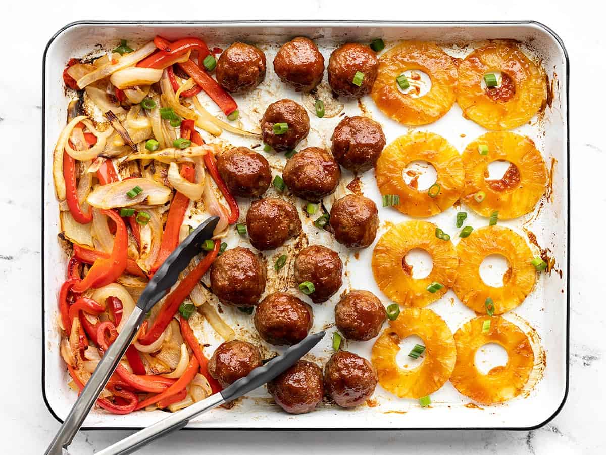 Overhead view of sheet pan with bbq meatballs, pineapple, peppers, and onions, tongs on the side