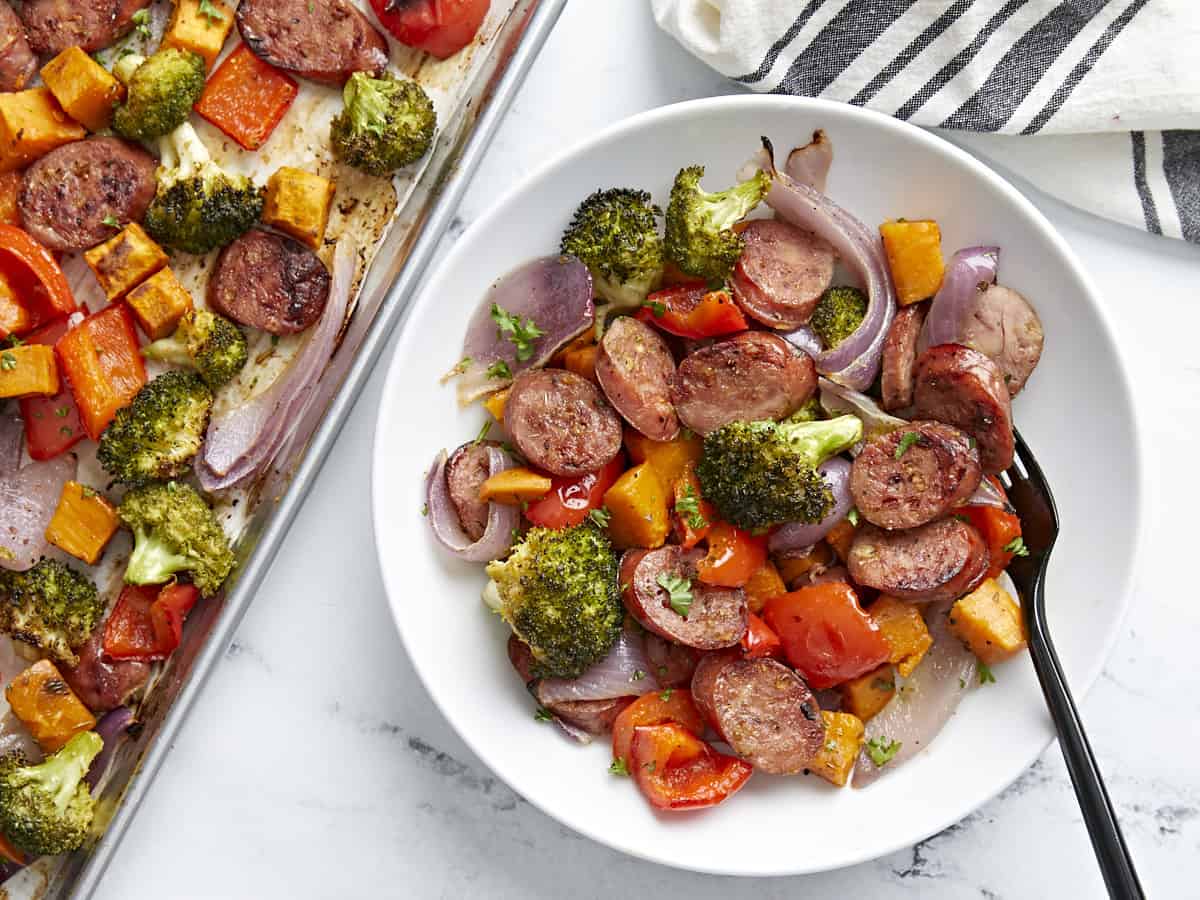 A serving of Sheet Pan Chicken Sausage and roasted vegetables on a white plate next to a sheet pan.