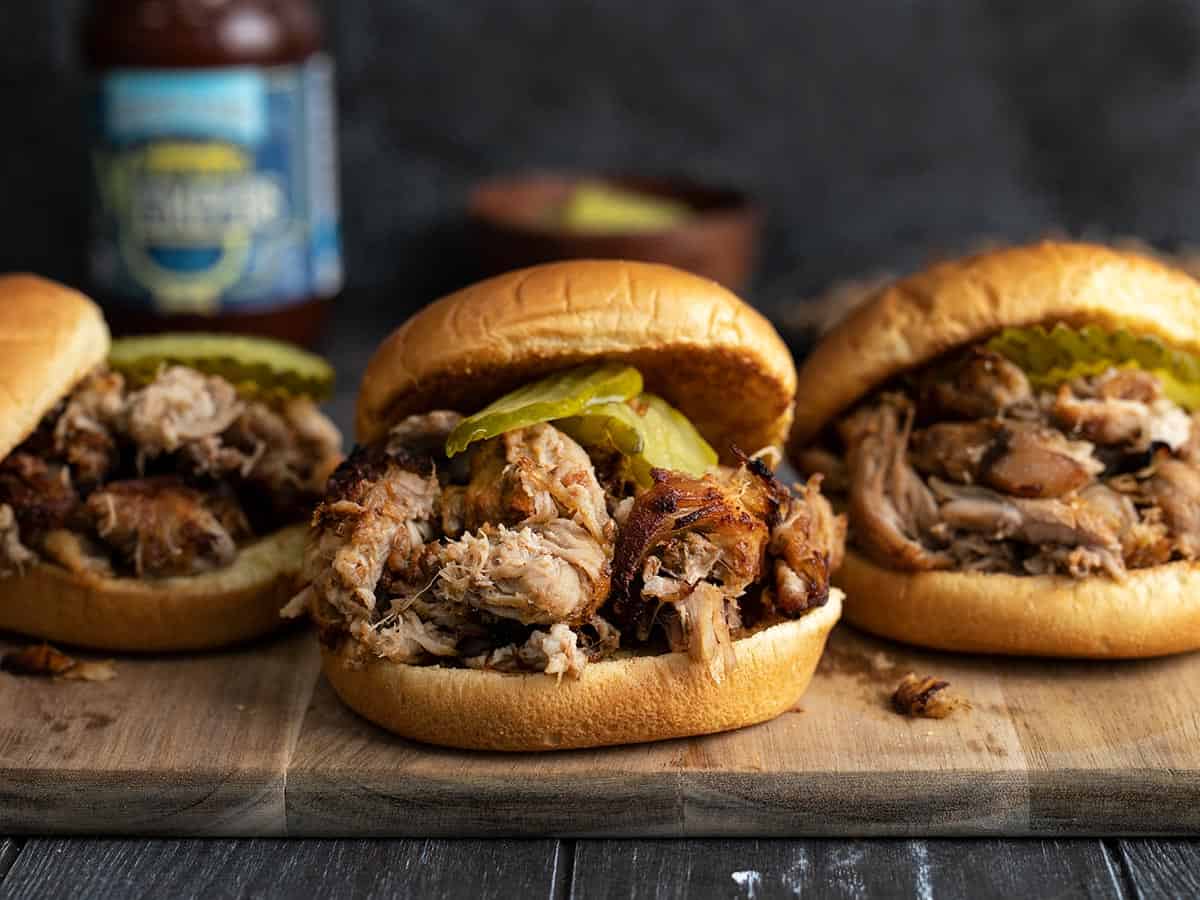 Three pulled pork sandwiches with pickles on a wooden cutting board.