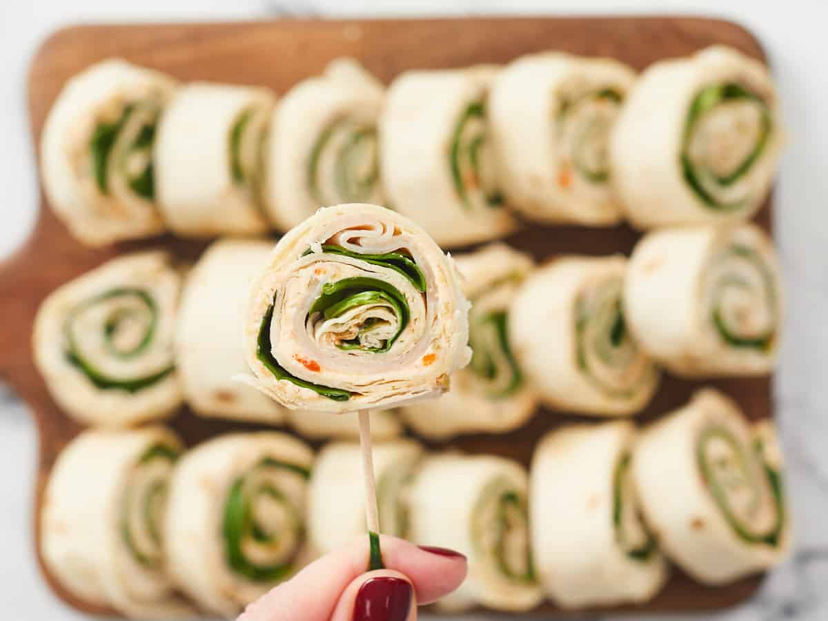 An extreme close up of one turkey pinwheel. In the background, there are many turkey pinwheels arranged in three rows on a wooden cutting board.