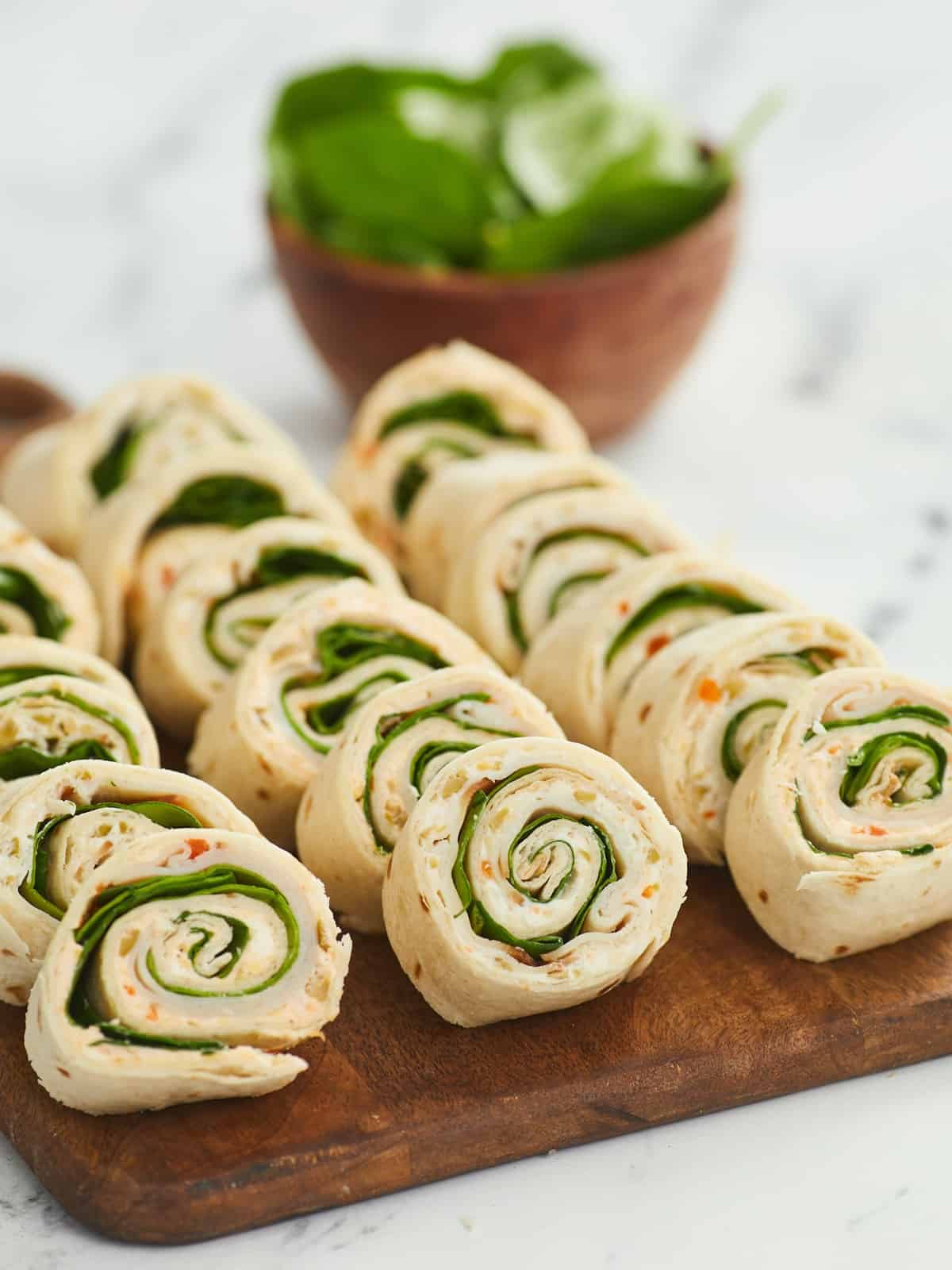 A close up shot of Turkey Pinwheels arranged in three rows on a wooden cutting board with a white background and a bowl of spinach blurred in the background