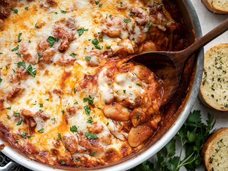 Close up view of a spoon scooping some white beans with mushrooms and marinara, garlic bread on the side.